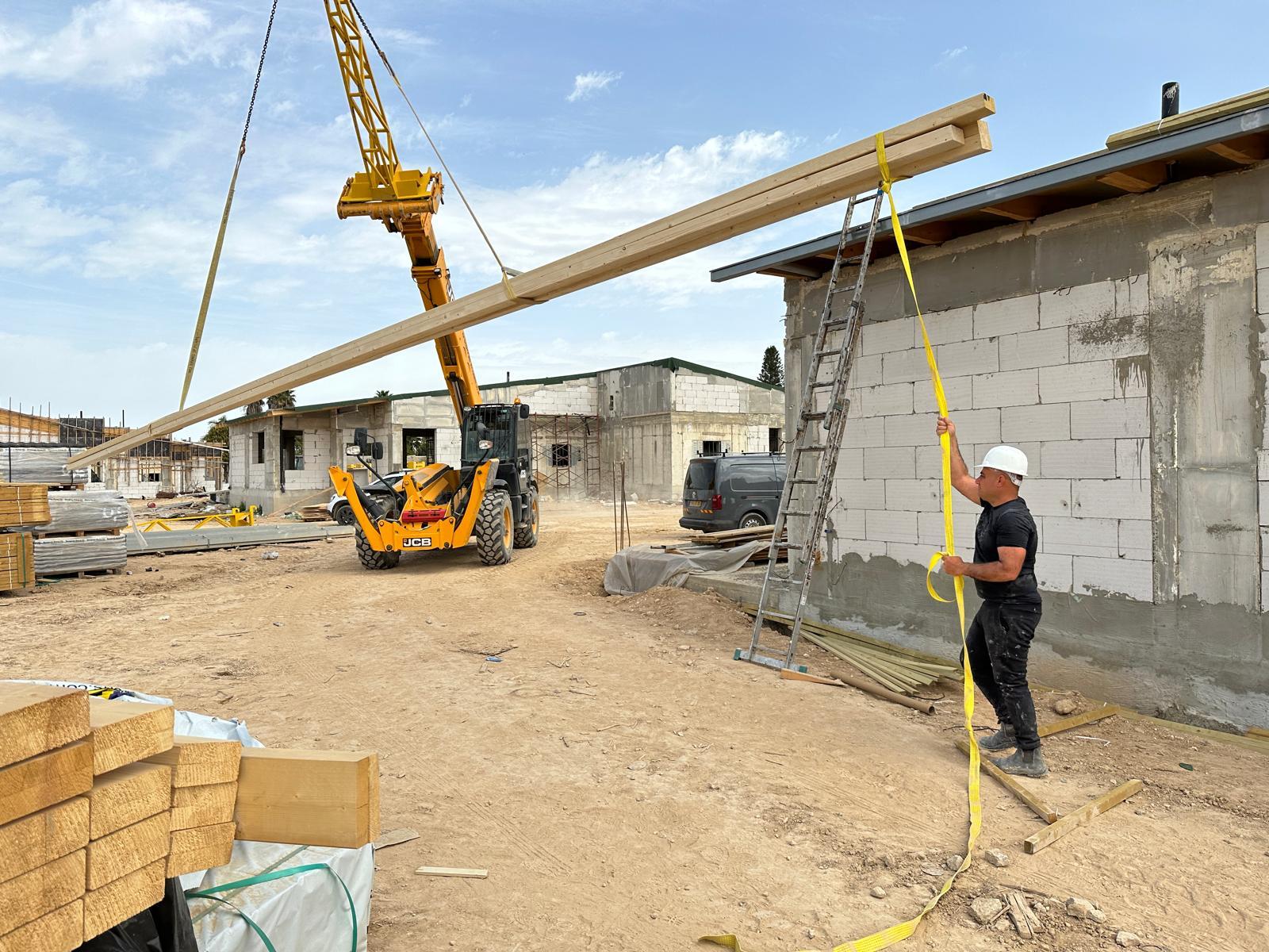 מנוף מרים קורות עץ לגג באתר בנייה בחצרים, עובד מכוון אותן בעזרת רצועה